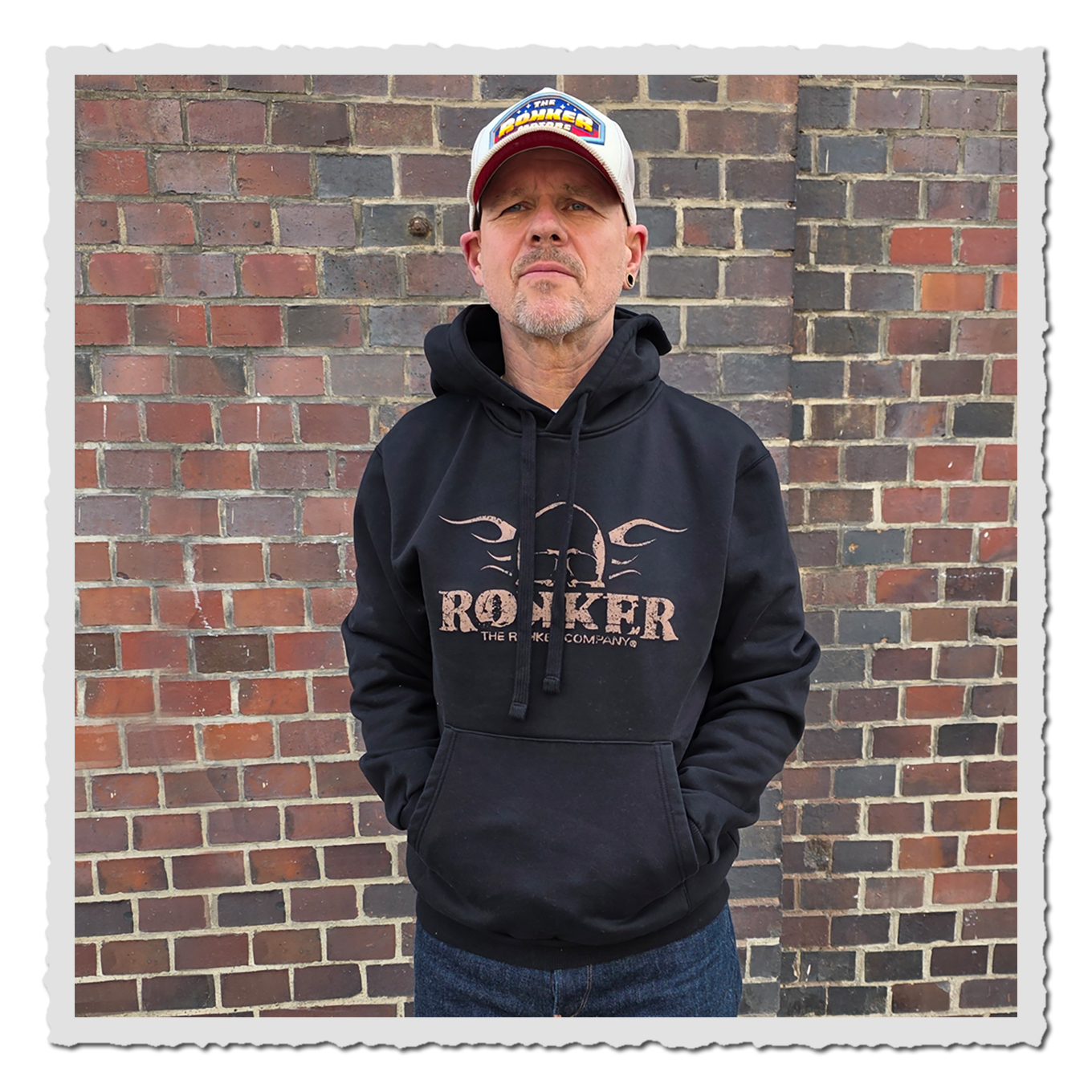 Person in schwarzem Hoodie mit Frontprint vor Backsteinwand, frontal fotografiert, mit Kappe und Jeans.